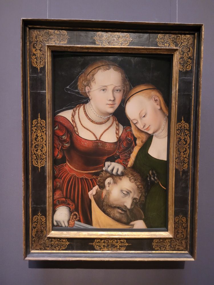 Ölgemälde in einem alten Holzrahmen, man sieht zwei Frauen, die den abgetrennten Kopf eines Mannes in der Hand halten, der gerade in einen Sack gesteckt wird. Judith trägt ein rotes Rennaisancekleid mit durchsichtigem Schleier am Kopf und hält ein Messer in der Hand. Sie hat den Blick nach vorne gerichtet und sieht entspannt aus. Ihre Gehilfin hat eine blonden langen Zoof und ein grünes Kleid. Sie blickt leicht lächelnd nach unten zum Stoffsack.