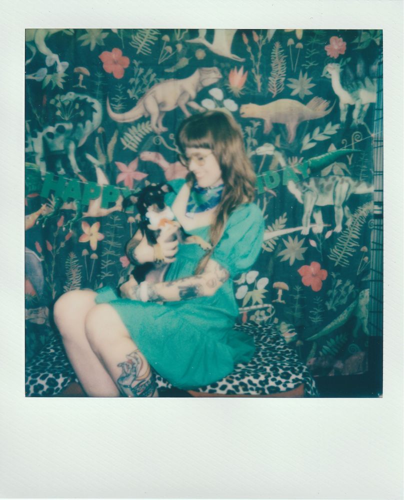 scan of a polaroid photo of avalon sitting on a leopard print blanket in front of a jungle dinosaur background with a banner that says “happy birthday.” They are wearing a green dress and holding their small black and tan dog Bardo at his 3rd birthday party. 