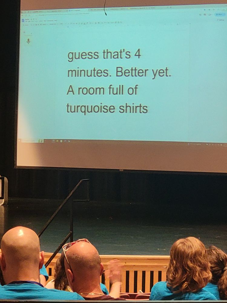 A screen on a stage showing voice tontext trqnscription that says "Better yet. A room full of turquoise shirts" with a room full of turquoise shirts in the foreground