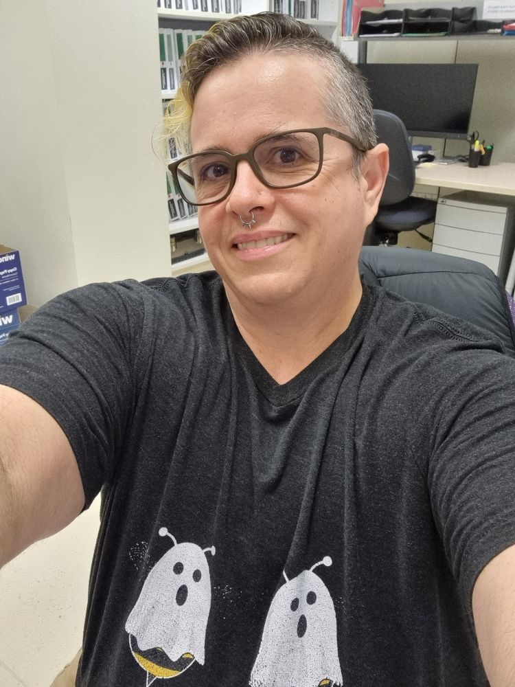 Selfie of Bee a white non binary person. 

They have short hair and a septum piercing.

They are wearing a black t-shirt that has two cartoon bees with white sheets over them, so they look like ghosts. 