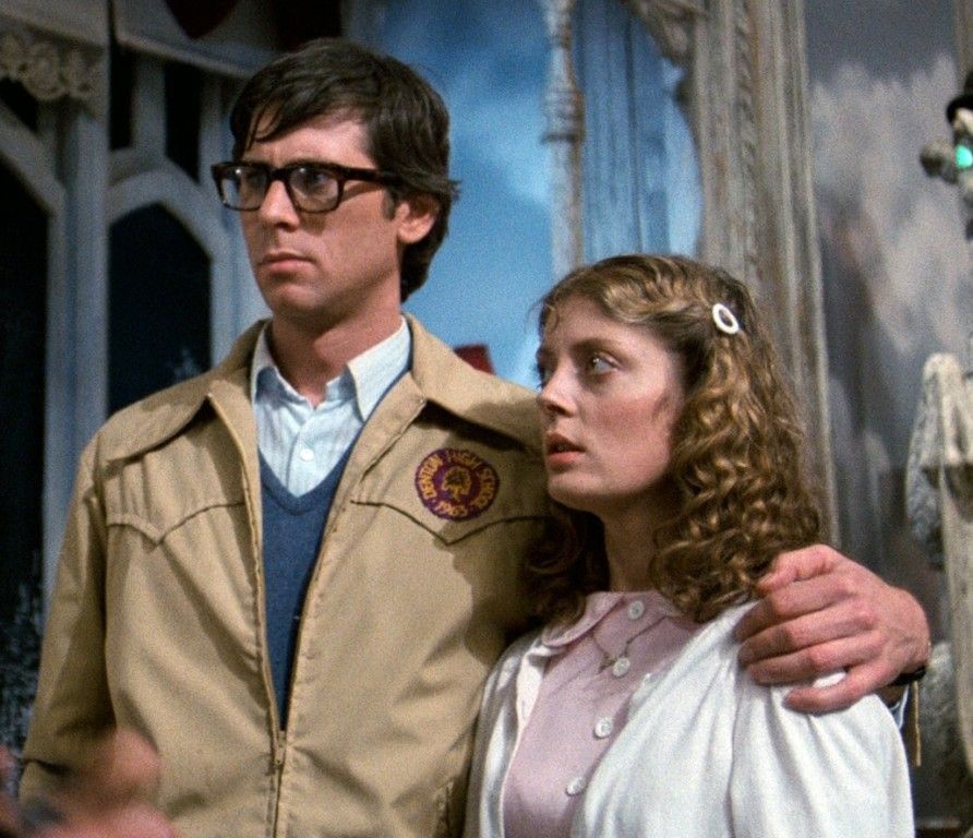 Still photo from the movie Rocky Horror Pictue show.

The image is from early in the movie and shows Brad with his arm around Janet.

Janet is a wholesome looking woman wearing a pink dress and a white cardigan.

Brad is a nerdy looking man wearing a button down shirt, vest, and a beige jacket. 