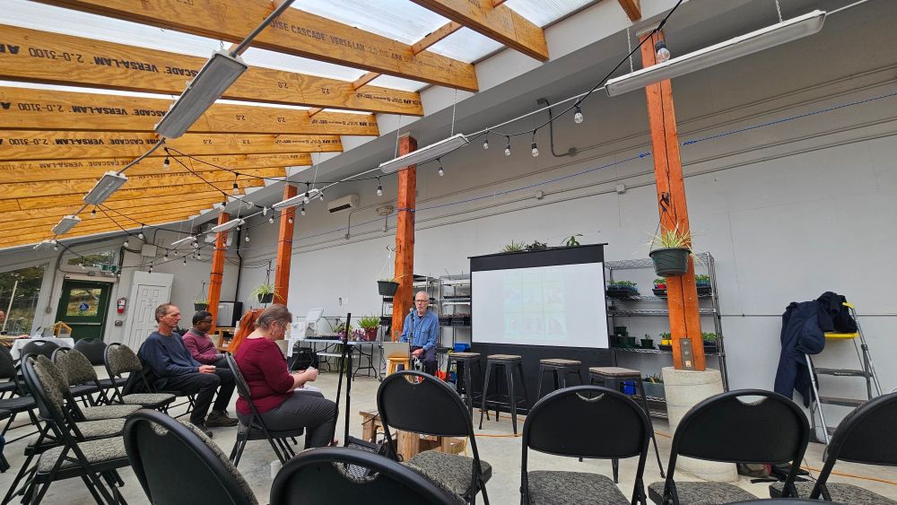 A man speaks to people on chairs in a greenhouse 