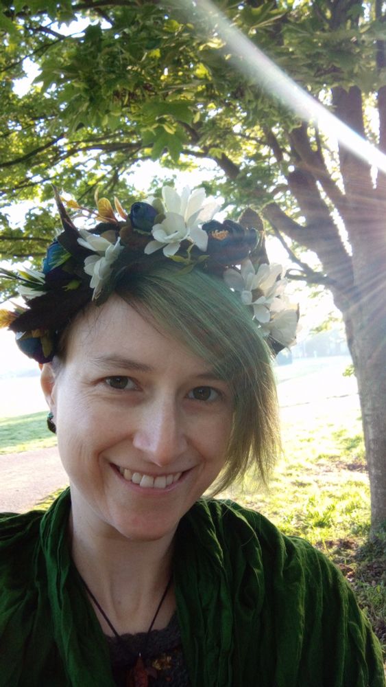 Roary, a white person with short hair and a longer green fringe on one side, smiles into the camera. They are wearing all green, with a flower crown of green, white and blue on their head. Behind them the early morning sun shines through the leaves of a tree.