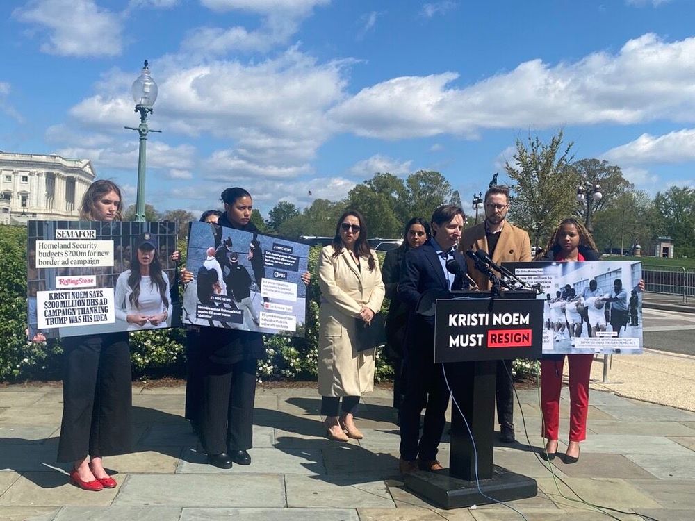 NIJC Senior Policy Jesse Franzblau speaks before a group of people standing behind a podium with signs, one reading "Kristi Noem Must Resign", during a press conference.
