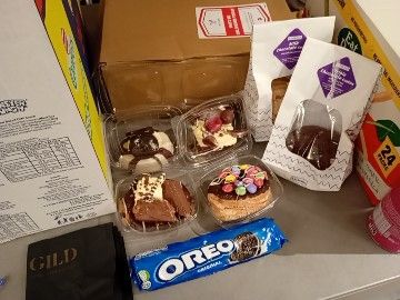 A selection of treats as described in the post laid upon a grey plastic table. The treats are surrounded by large cardboard boxes most likely full of food brought by other attendees. This demonstrates the meager nature of my offering.