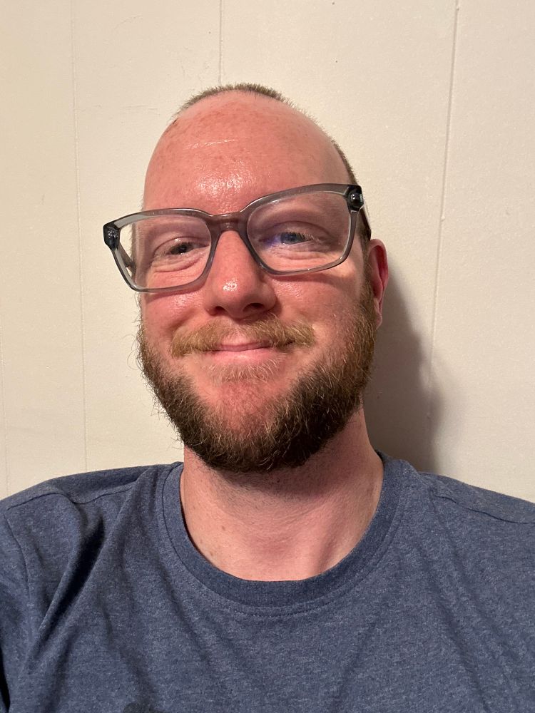 A selfie of me smiling and sitting against a white-ish wall. I’m wearing a blue shirt.