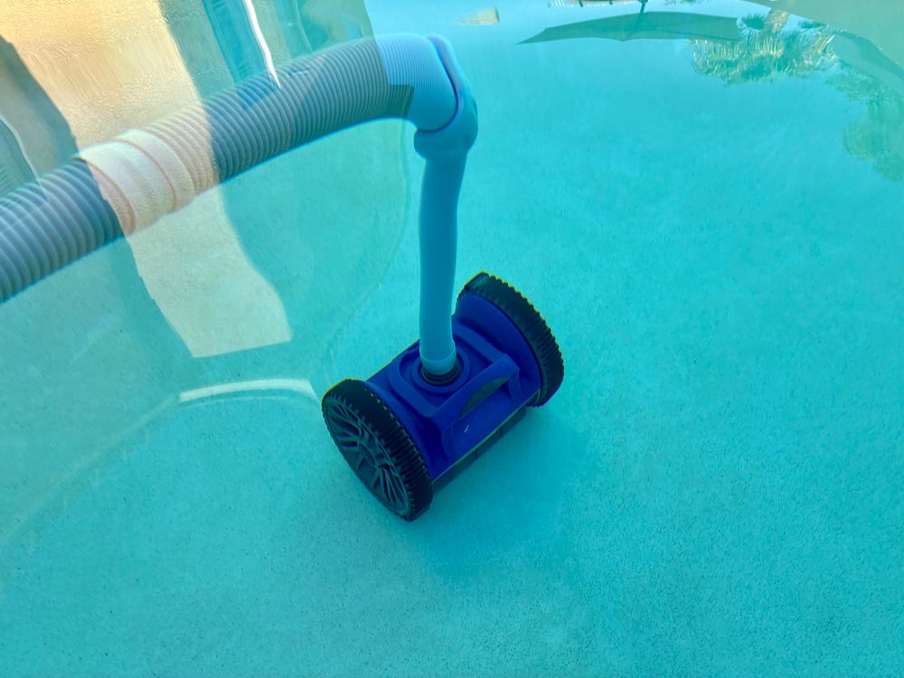 Photograph of a swimming pool sweeper under the still water surface. It is royal blue with two large black wheels, and a grey hose.