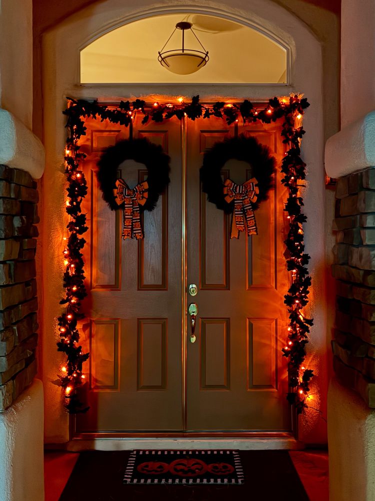 Photograph of a double door entry decorated with black Halloween wreaths and garlands, orange lights, and matching bows/doormat. 