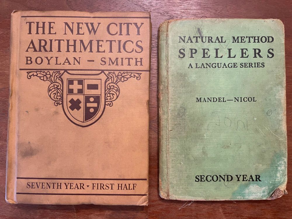The two books, on a wood surface. The arithmetic book is larger, brown, and in good condition. The spelling book is green, smaller, and in less good condition.
