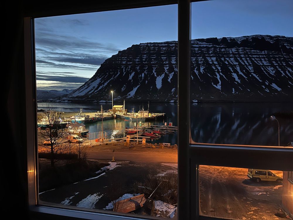 Morning view out of a window of a small fishing harbour with lights on with a Fjord bluff behind it