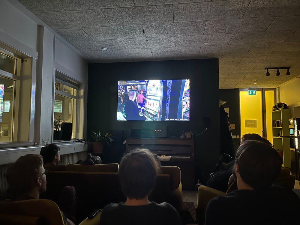 Screen showing a still from Grand Theft Hamlet (people in a casino playing slot machines) from the perspective of an audience member in communal space