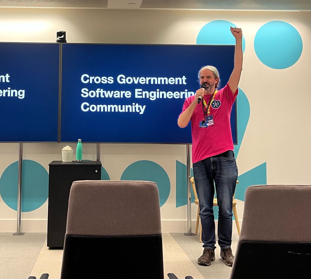 A man in pink shirt punches air in front of screen that reads - Cross Government Software Engineering Community