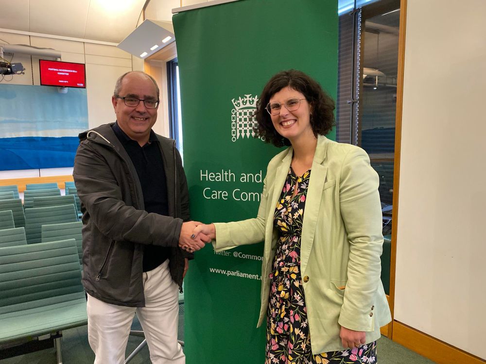 A man and a woman stand shaking hands in front of a green banner