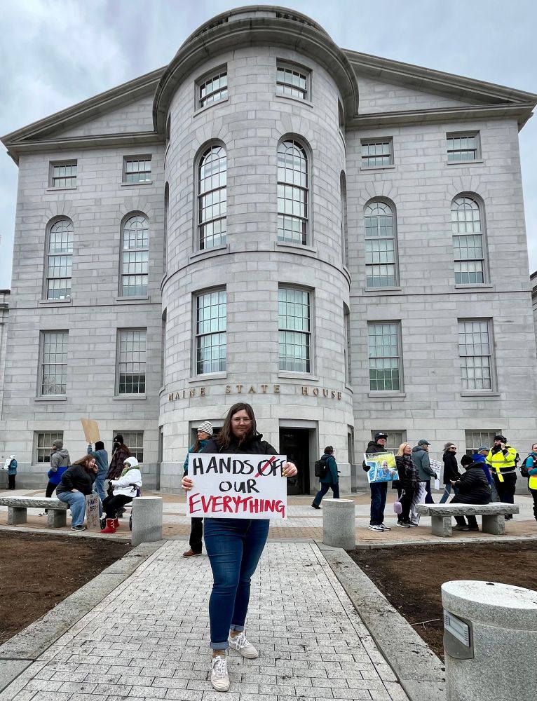 Woman standing in front of Maine State House with a sign that says hands off our everything 