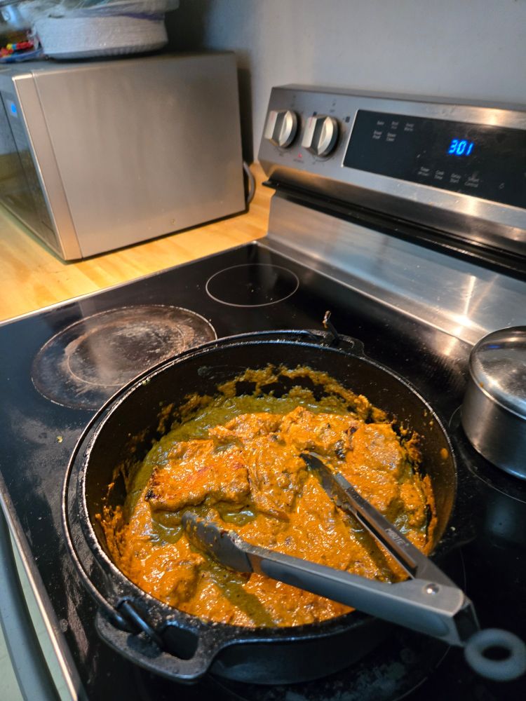 A deep cast iron Dutch oven on a stove, filled with butter chicken and sauce, with a pair of tongs resting on top.