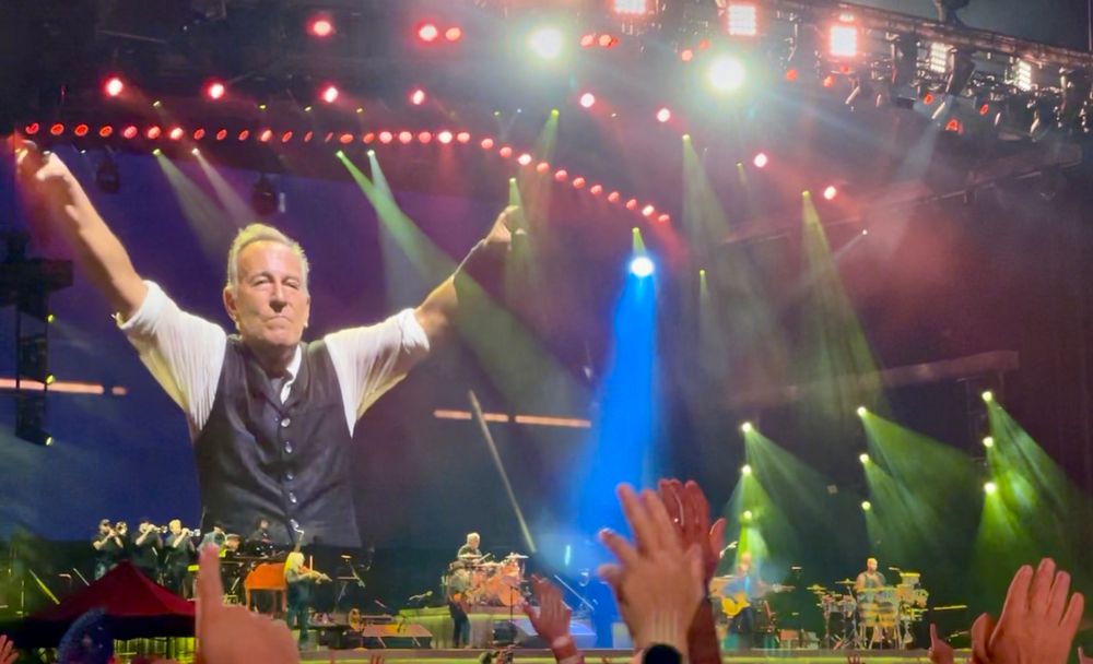 At a Bruce Springsteen concert in San Sebastían, Spain. 🇪🇸 The Boss is on the giant screen with his arms outstretched. Flashing lights all around. Hands are up in the air in the foreground. 