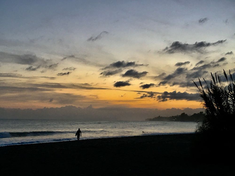 A lone surfer walking on the beach with the last light of the day. The surfer can be seen in silhouette. The sky is awash with the last colors of the sunset. 