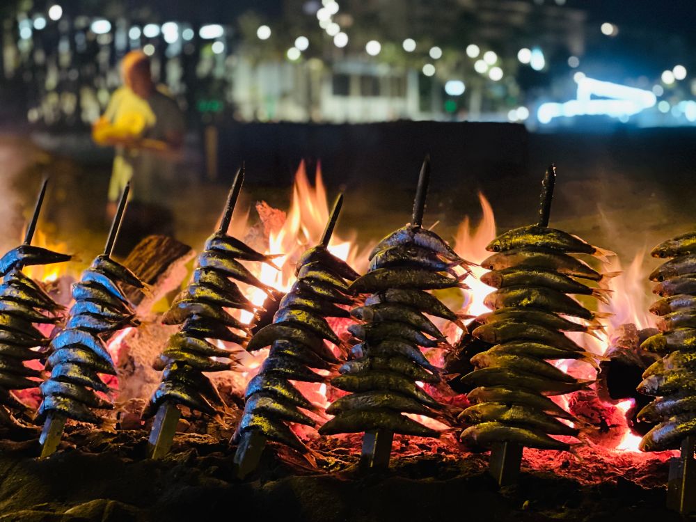 Sardines on a spit coking over a wood fire on the beach. The beach is behind the sand filled grill which is in an old fishing boat. The town lights off in the distance. 

