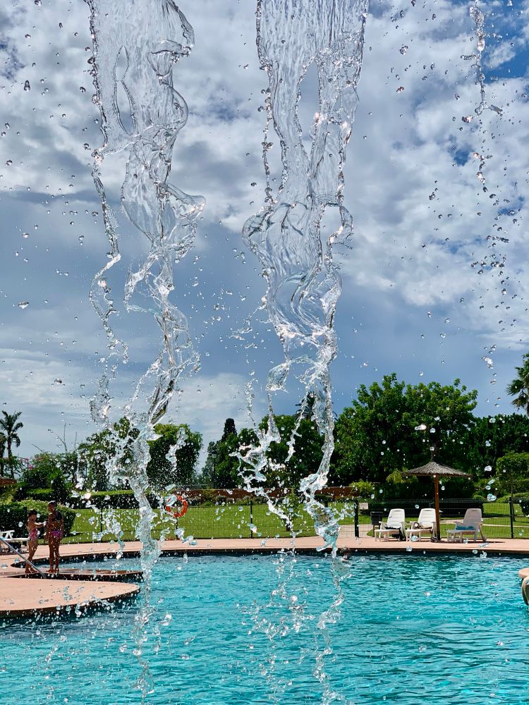 Under a waterfall. The falling water is in the foreground. In the background is a large pool with chairs and umbrellas. A couple of kids are playing. The garden is beyond that and the sky has some large clouds floating across it. 