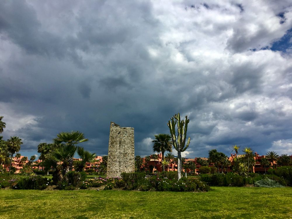 Storm clouds are brewing. Underneath the angry sky is an ancient lookout tower which is surrounded by palm trees, bushes and flowers. 