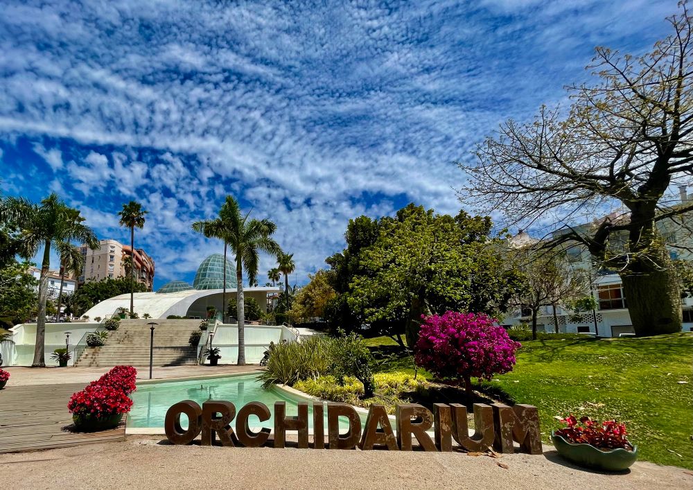 In the foreground in large letters it says “Orchidarium”. It is the largest Orchid house in Europe. Behind the letters is a grassy area with blooming bougainvillea and some trees on the right. A pool and walkway on the left. Behind those the orchid house can be seen. A large glass dome surrounded by palm trees. Behind it some apartment buildings are sticking out. The sky is covered in wispy clouds. 