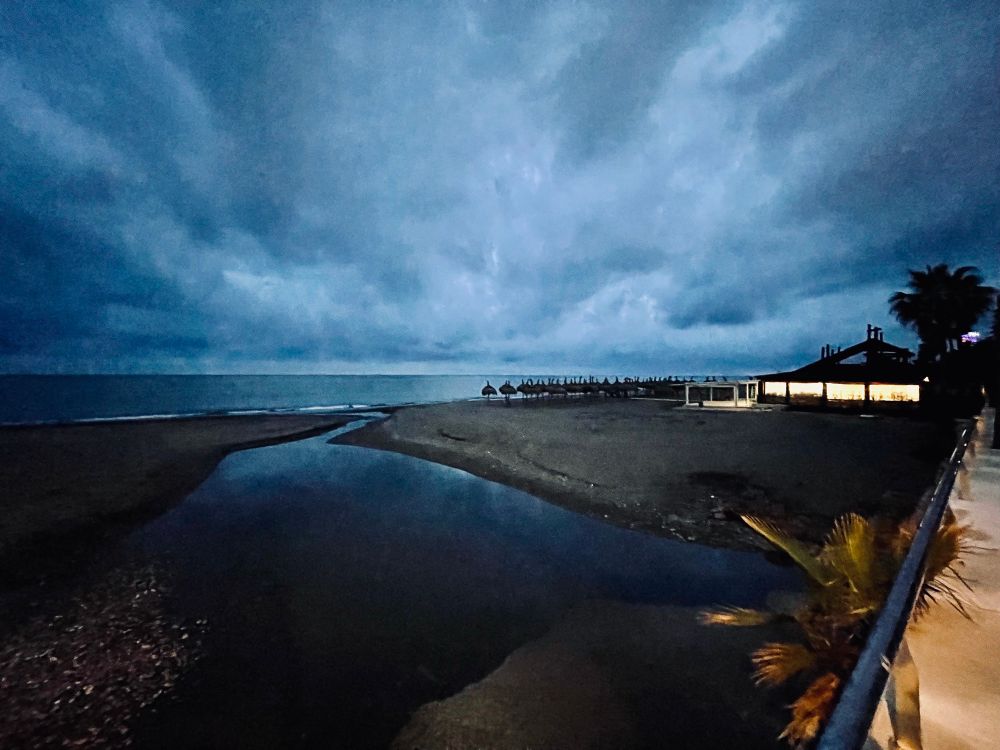 Storm clouds have gathered. A river runs into the sea and a Chiringito is lit up for the evening crowds. 