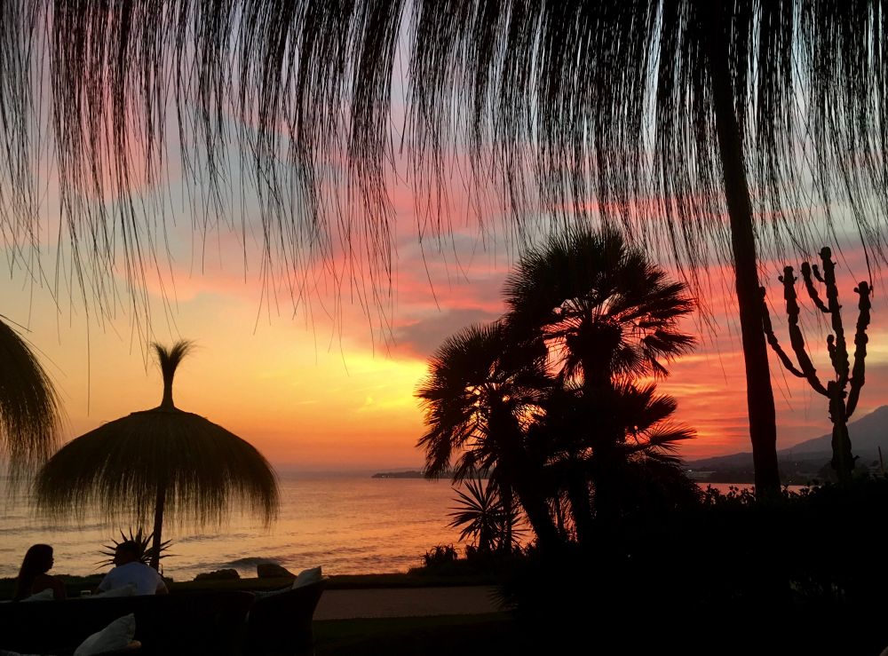 Sunset on the beach. The picture is taken from underneath a straw umbrella. Another umbrella can be seen on the left side with people talking and plants on the right, all in silhouette. The sky is bright with sunset colors from yellow to dark orange. The sea is reflecting some of the sunset colors. 