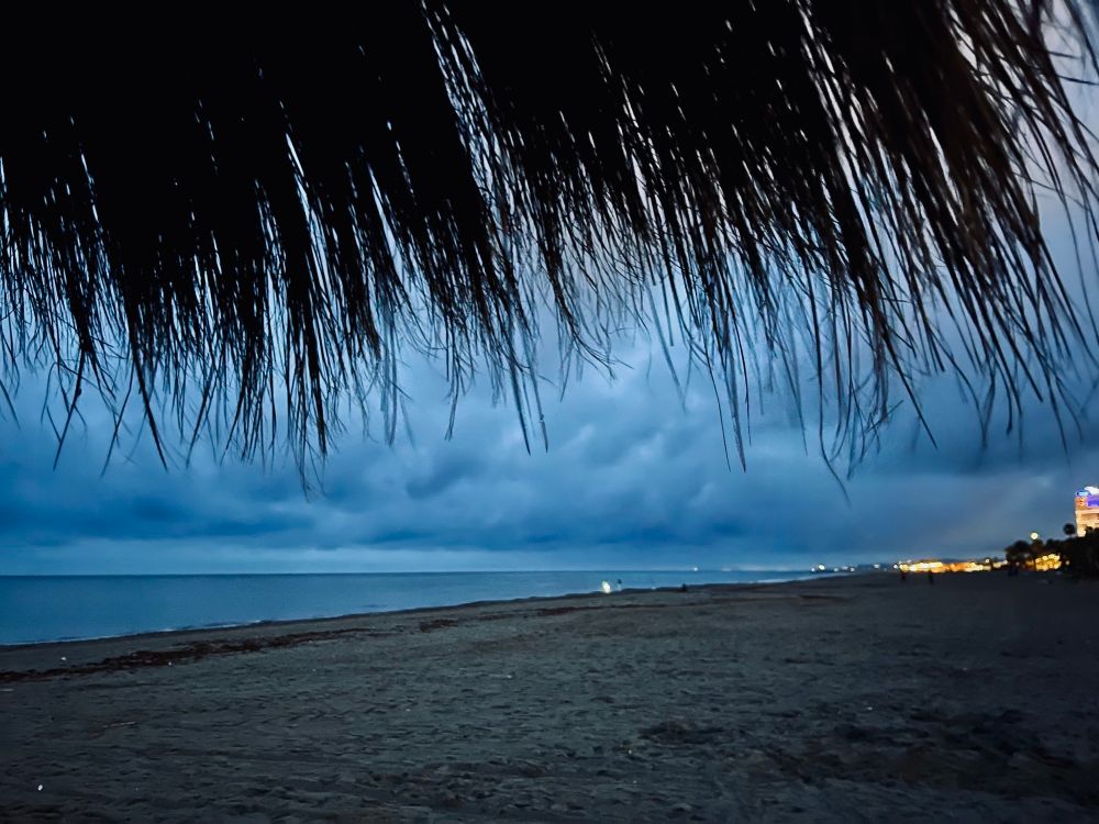 On the beach looking towards the harbor. On the right some town lights can be seen. On the left and center the sea. The storm clouds have a dark blue glow to them. The top of the picture is fronds from an umbrella. 