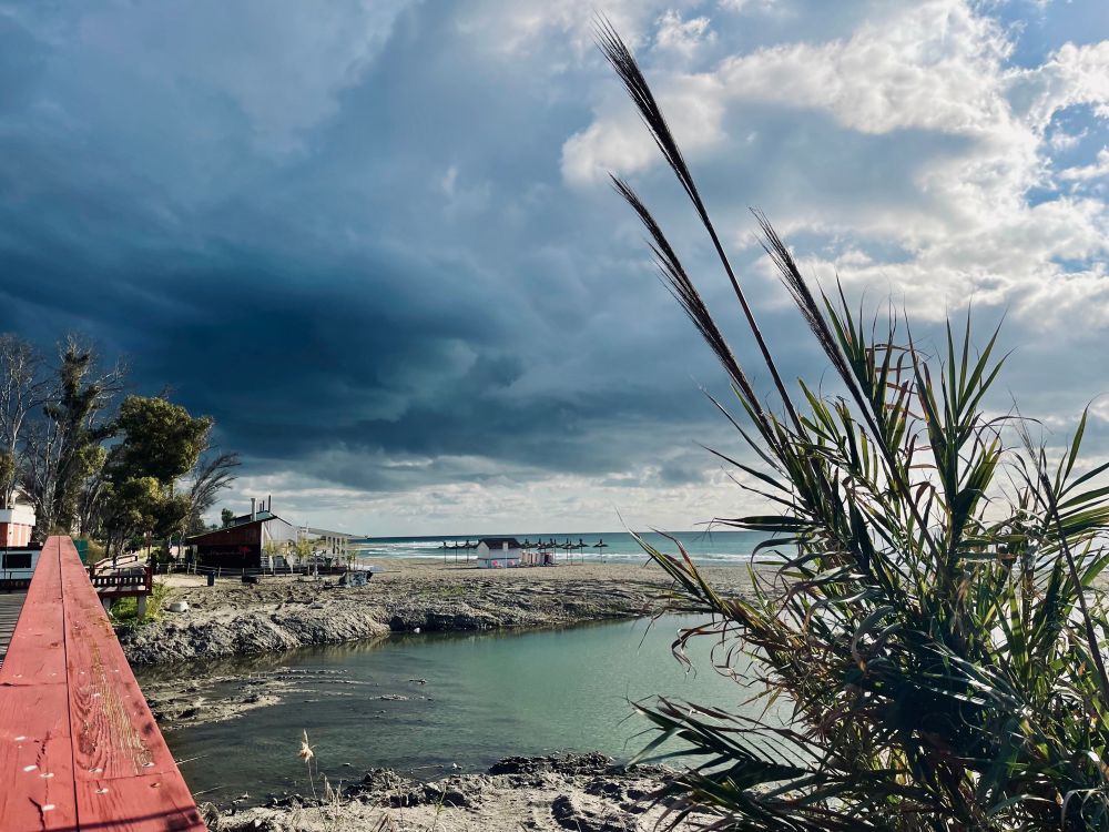 Walking along the board walk next to the Mediterranean Sea. The storm clouds are gathering and looking very ominous in the not so far off distance. 

Some reeds are on the right. A river runs into the sea in front of us with the beach behind it. The med is just past the sand and a Chiringito under some ancient trees is on the left. 

Those storm clouds did catch up to me before I could find shelter. After a good downpour and some rather wet clothes they moved on to their next destination. 