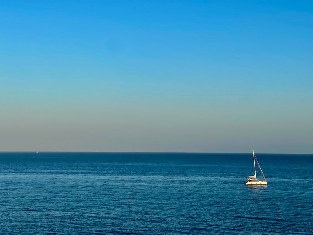 A minimalist photo of the Mediterranean Sea taken in Estepona, Spain. The Med is very calm and a catamaran is lazily bobbing in the water. There is not a cloud to be seen. The haze is changing to sunset colors ever so gently. 