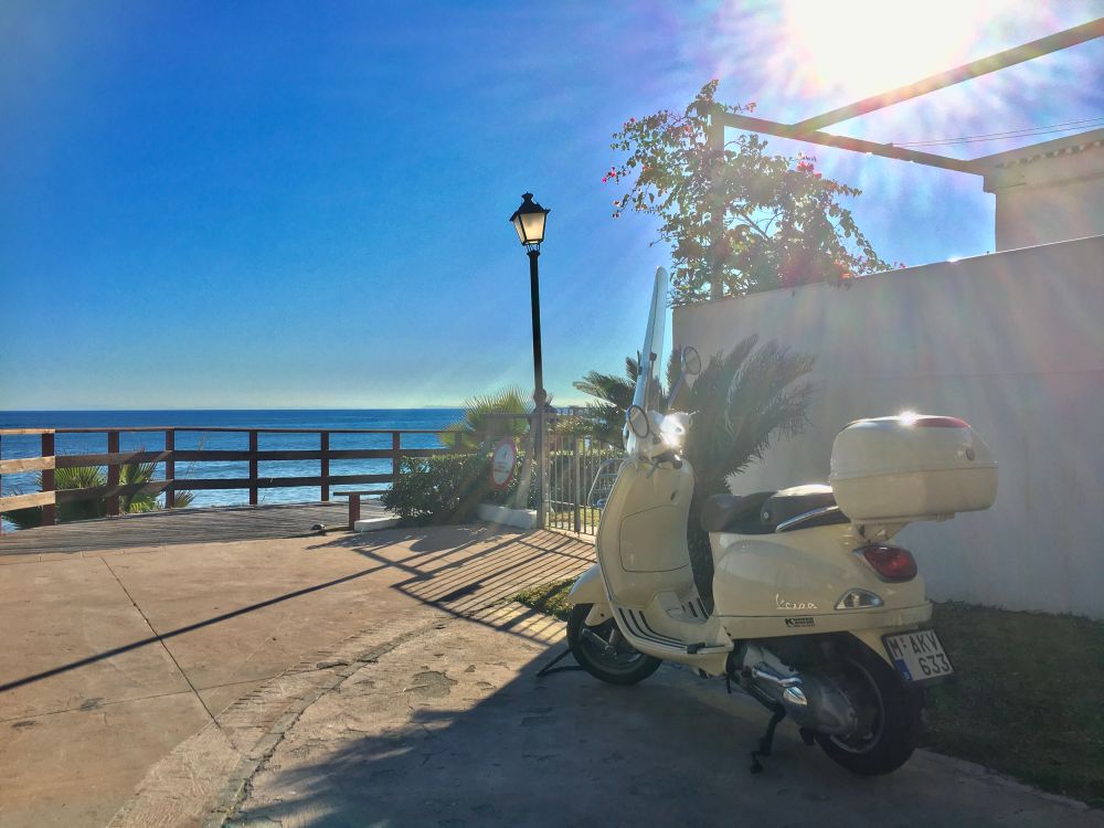 A Vespa sits idle by the seafront. Bougainvillea growing over a wall that cannot contain them. A lantern lamp is being lit up by the brilliant sunlight. The sea is calm. 