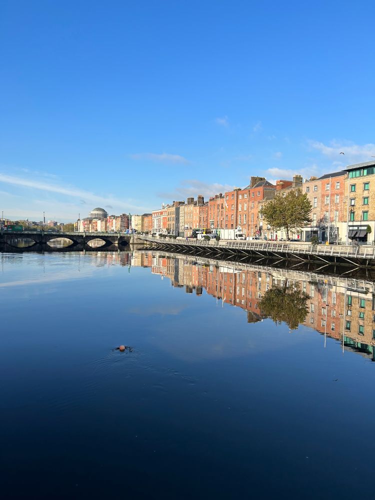 View of Dublin up the Liffey from the Millennium Bridge. Clear blue sky and deep blue water. Some of Four Courts visible in the distance. Buildings on the northside reflected clearly in the river.