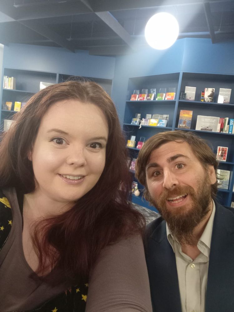 Dr Krysia Waldock is on the left, and Dr Nathan Keates is on the right, and behind them is a blue library with various books on the shelves. 