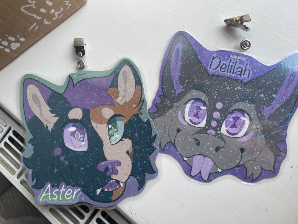Two laminated, holographic fursuit badges on a white windowsill. One badge is a black, orange, and purple cat smiling with the name Aster. The other is a purple and grey dutch angel dragon sticking it’s tongue out, with the name Delilah.