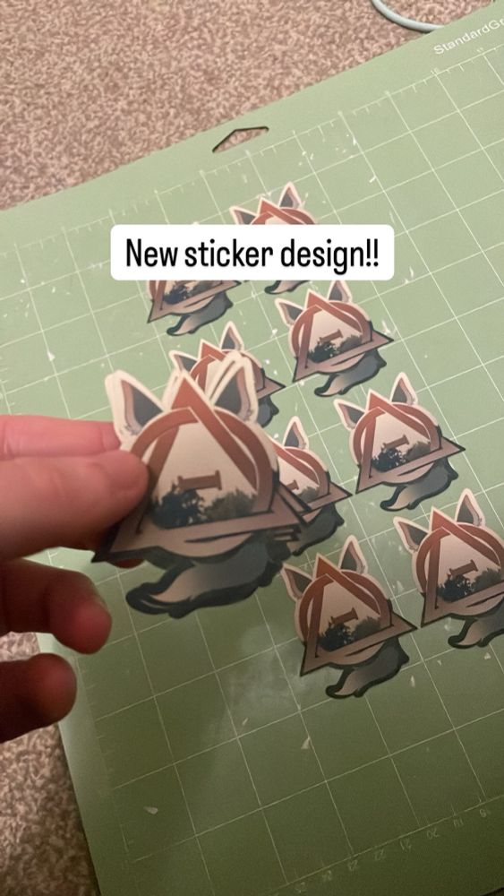 A photo showing a hand holding a sticker of the therian sticker with animal ears and a tail, and a forest background. Several others of the sticker are behind the hand on a craft mat. Text says ‘new sticker design!!’