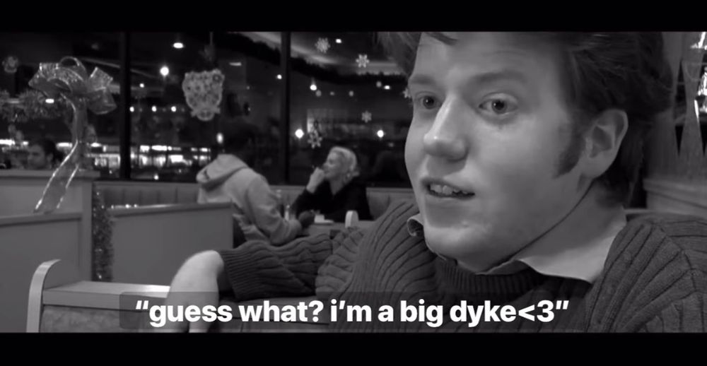 a screenshot of brennan lee mulligan, looking very young, sitting in a diner with a suit on. the subtitles show him saying "guess what? i'm a big dyke"