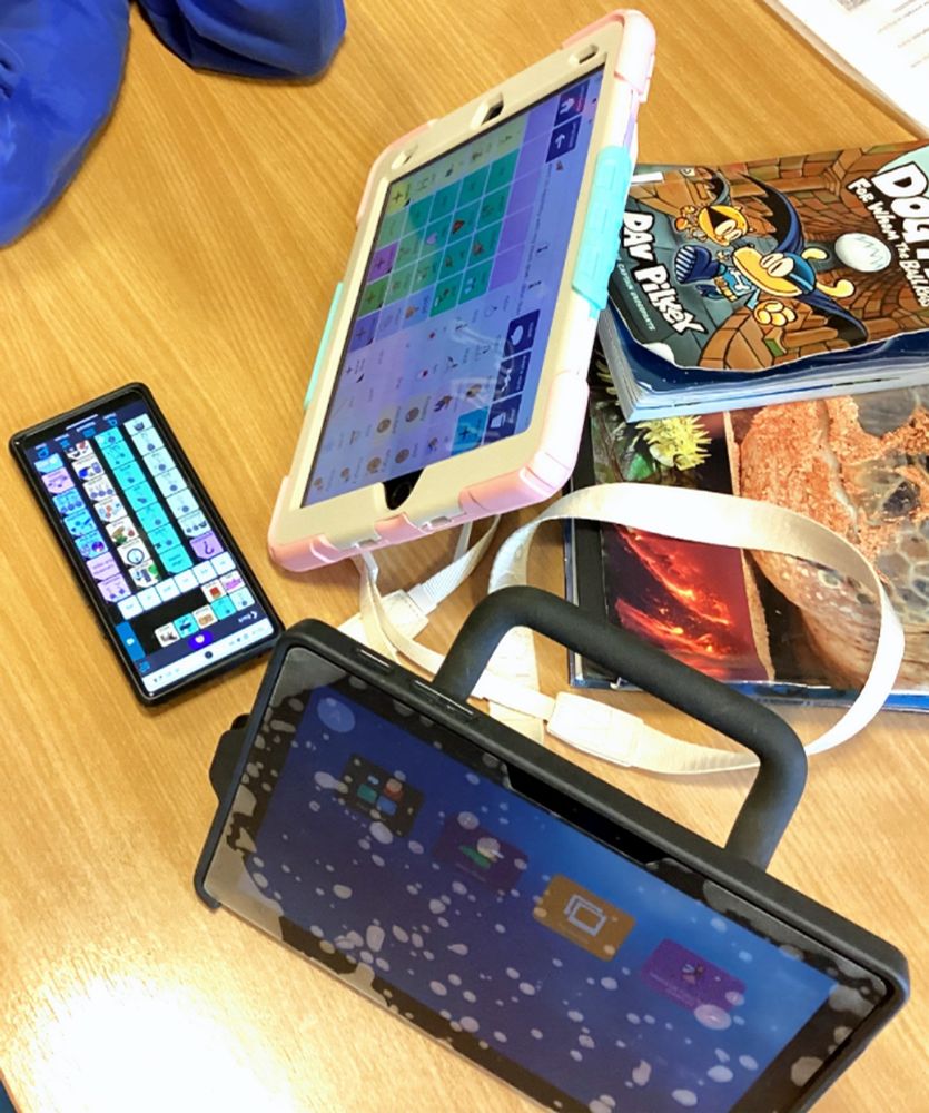 Photo shows a table, on top of which is a mobile phone with the WeaveChat communication app's scrolling board, an iPad with Smartbox's Grid SuperCore 50 app and a Smartbox Touch Pad displaying the Grid dashboard. There are some books on the table behind the devices.
