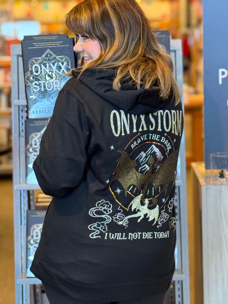 Girl with a black sweater that reads Onyx Storm, Brave the dark, I will not die today, with golden dragons on it. In front of Onyx Storm book shelf
