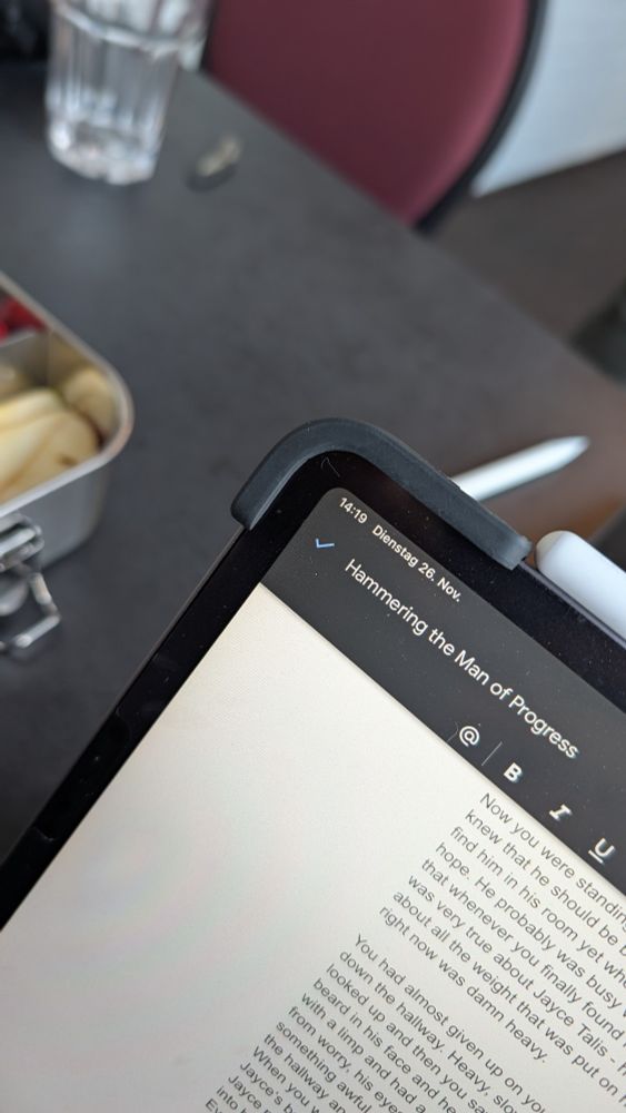 The corner of an iPad pro 11" in the foreground with Google docs open. The file name is "Hammering the Man of Progress". In the background you can see a bit of a lunchbox with apple slices in it on a grey table. There's also a glass and an apple pen. 