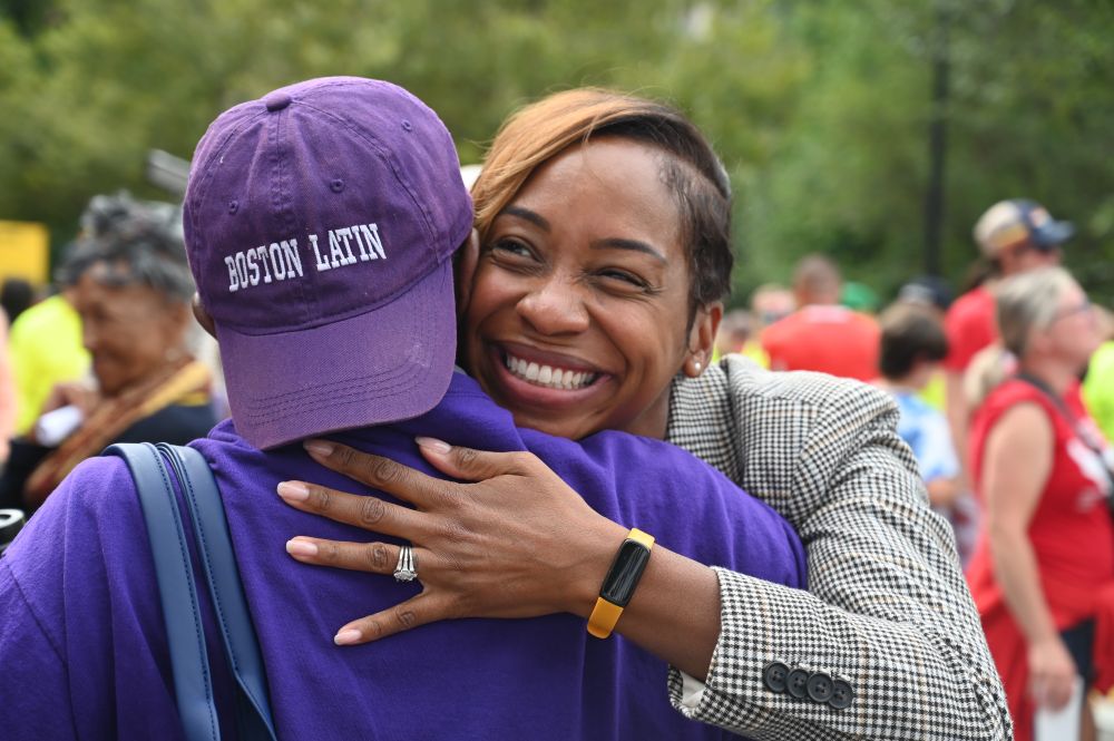A photo of AG Andrea Joy Campbell hugging a person in a purple shirt with a purple hat that says 