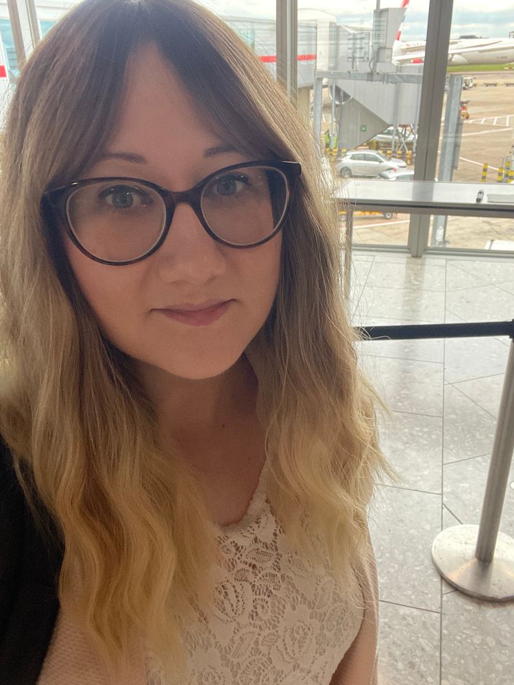 Sulin, a white blonde woman with wavy hair and a white lace top taking a selfie in an airport. Behind her you can see the wing of a plane out the window.