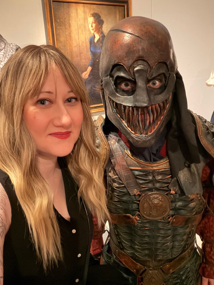 Sulin, a white blonde woman with wavy hair and a black sleeveless shirt, takes a selfie with a Seanchan soldier in full costume.