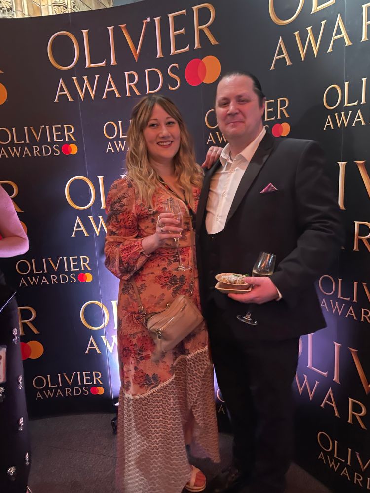 Sulin, with blonde mid length curly hair in a long long floral dress holding a margarita, with Rob, with dark ties back hair, in a tuxedo. They are standing in front of a billboard with the Olivier awards logo.