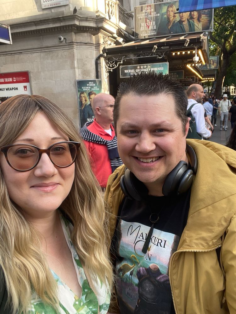 Sulin, a blonde woman with sunglasses and a green and white dress, taking a selfie with Rob, who has short black hair, a black MalkieriCon t shirt and yellow coat. They are in front of Wyndhams Theatre where you can see a billboard advertising My Master Builder.