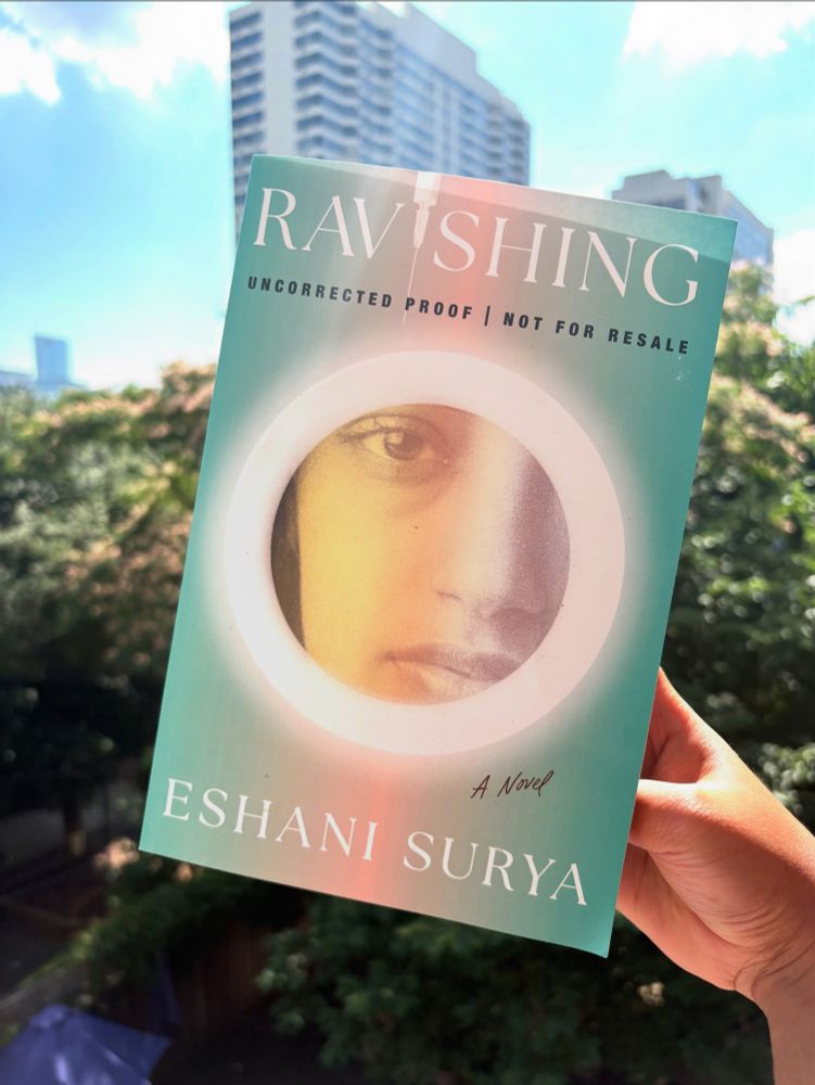 A photo of my book, RAVISHING, held up in front of a tree and a cityscape 