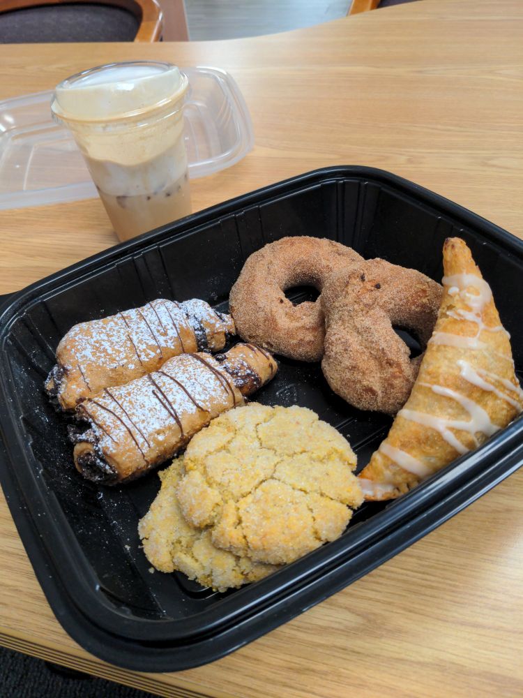 Black reusable take-out box with vegan apple turnover, cinnamon sugar donuts, cornmeal cookies, chocolate cigars(!) and pumpkin pie spice chai latte with cold foam. The latte is half full because of deliciousness. Clear plastic lid is in the background. All set on a generic office desktop.