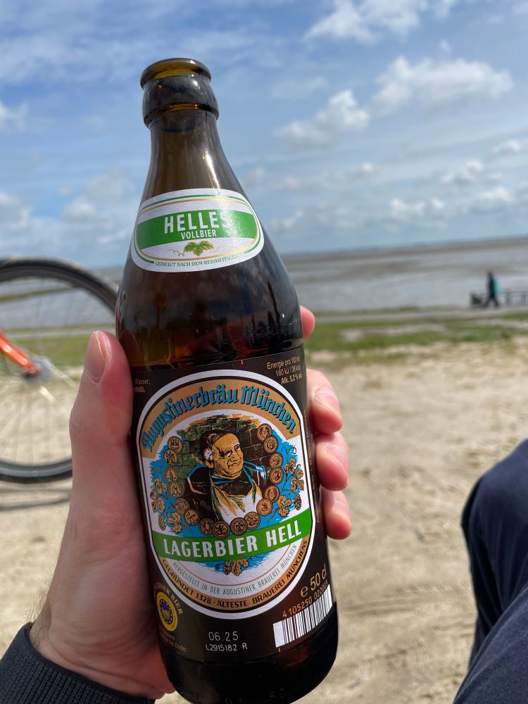 Eine Hand hält eine Flasche „Augustiner Bräu München Lagerbier Hell“ vor einem Strand mit Meerblick. Im Hintergrund sieht man einen Teil eines Fahrrads, eine sitzende Person und einen teils bewölkten Himmel. Auf dem Flaschenetikett ist ein bayerischer Mönch mit Münzen um ihn herum abgebildet.