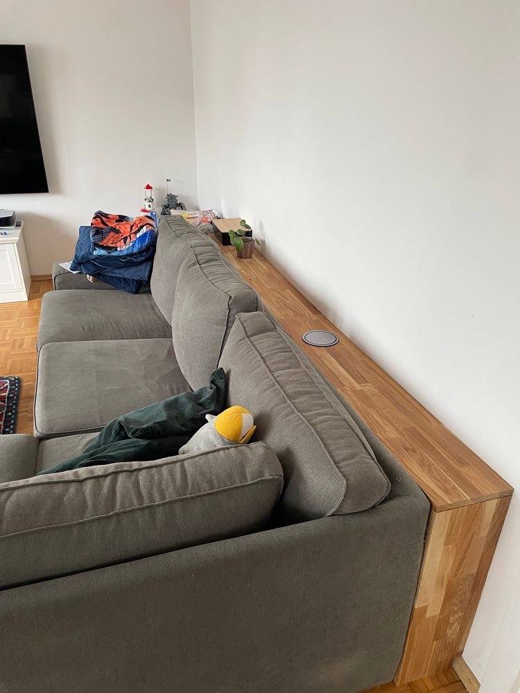Ein Wohnzimmer mit einem großen, grauen Sofa, das mit dem Rücken zu einer weißen Wand steht. Hinter dem Sofa befindet sich ein maßgefertigtes Holzregal, das die gesamte Länge der Wand einnimmt. Auf dem Regal steht eine kleine Pflanze und ein paar Gegenstände. Auf dem Sofa liegen eine grüne Kopfkissen und ein gelb-weißes Kuscheltier. In der Ecke ist ein Stapel bunter Decken zu sehen. Links hängt ein großer Flachbildfernseher an der Wand. 
