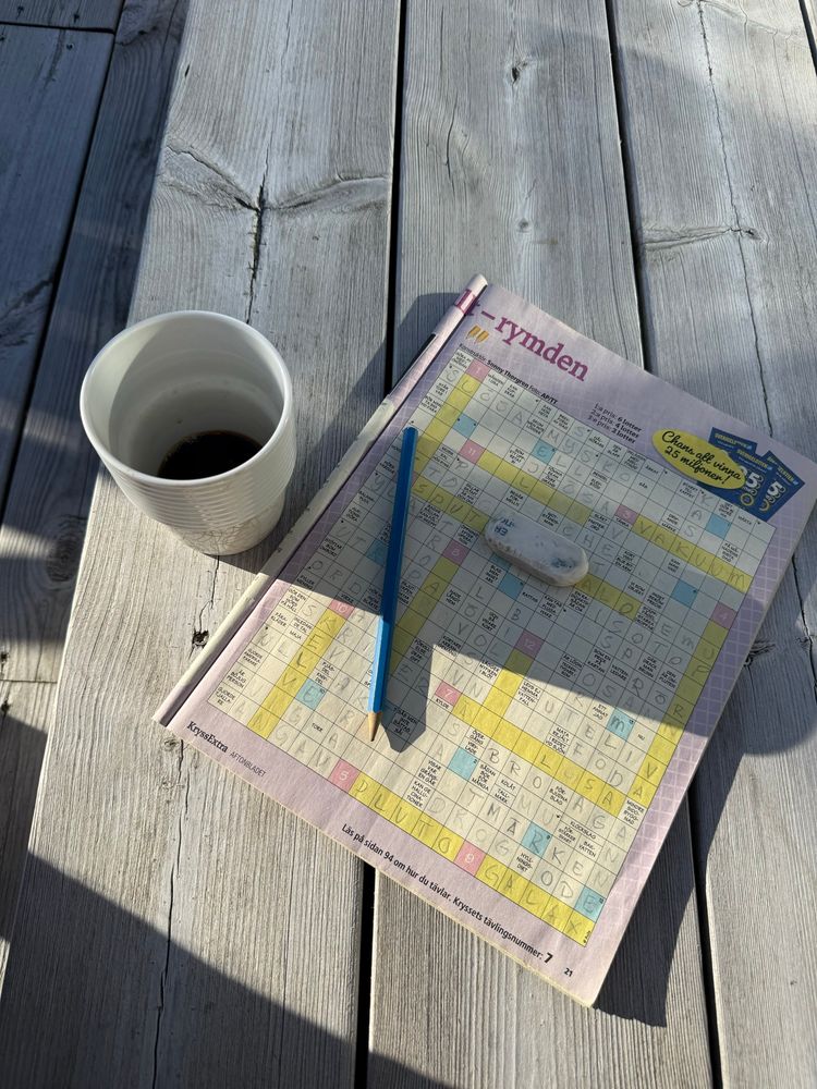 En kopp kaffe, ett korsord, en penna och ett sudd. På ett träutedäck i morgonsolen