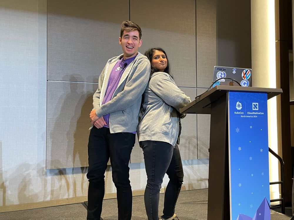 Picture of Leigh & Pinky posing back to back with their arms crossed in Flux hoodies at the podium on stage at KubeCon 2024.